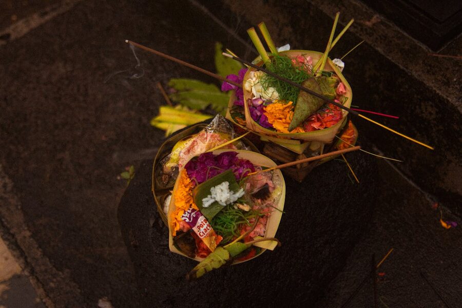 Two canang sari offerings stacked on a stone shrine in Bali, with incense smoking and flowers, rice and a small cracker on top