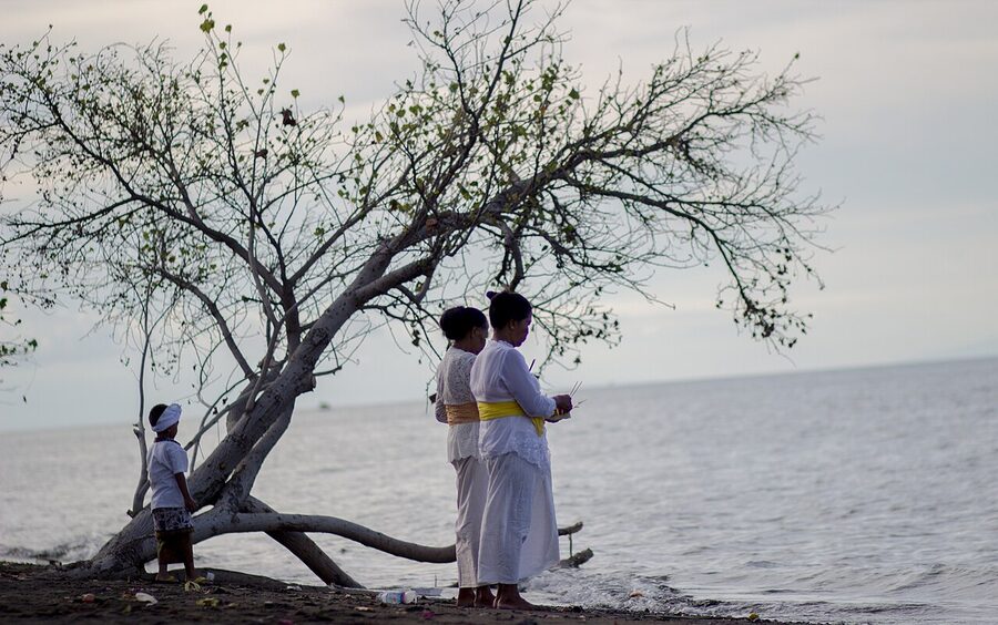 Three people in white ceremonial Balinese dress standing on the shore at Umeanyar Beach during a Melasti procession in north Bali