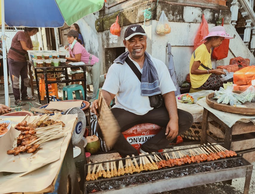 A smiling Indonesian street vendor grilling sate skewers over hot coals at a Bali market stall