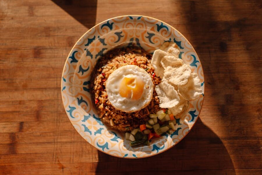 A patterned bowl of nasi goreng topped with a sunny side up egg, prawn crackers and pickled vegetables, viewed from directly above