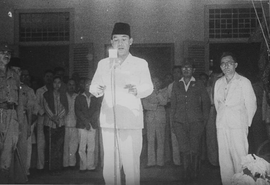 Black and white photograph of Sukarno at a microphone reading the Indonesian proclamation of independence in 1945, surrounded by associates