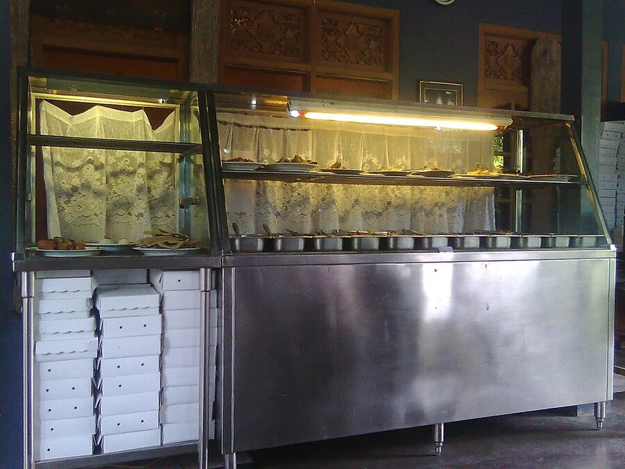 A glass-fronted warung counter in Bali with rows of metal trays of Indonesian food kept warm under a fluorescent light