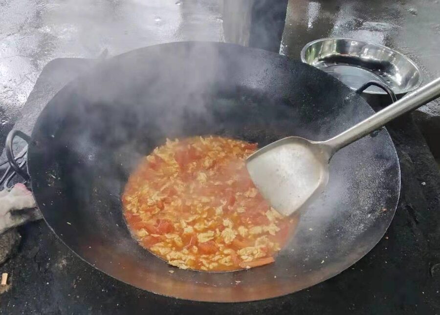 Tomato and egg sizzling in a black carbon steel wok over an outdoor stove with steam rising