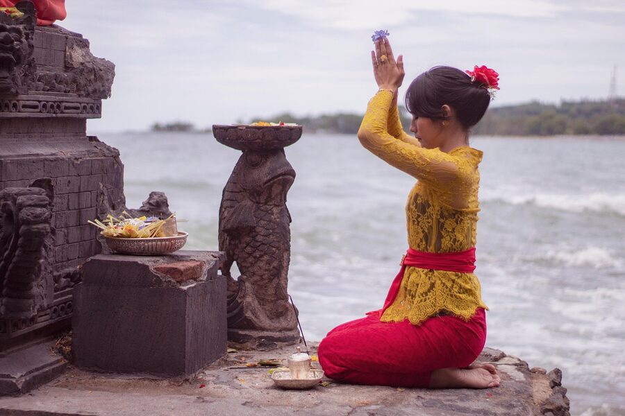 Balinese woman in yellow kebaya praying by the sea with offerings on a stone platform