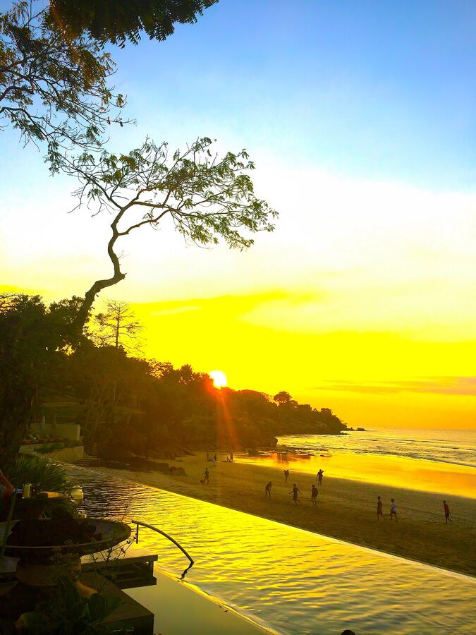 Jimbaran Bay at sunset with the silhouette of a tree and people on the wide flat beach
